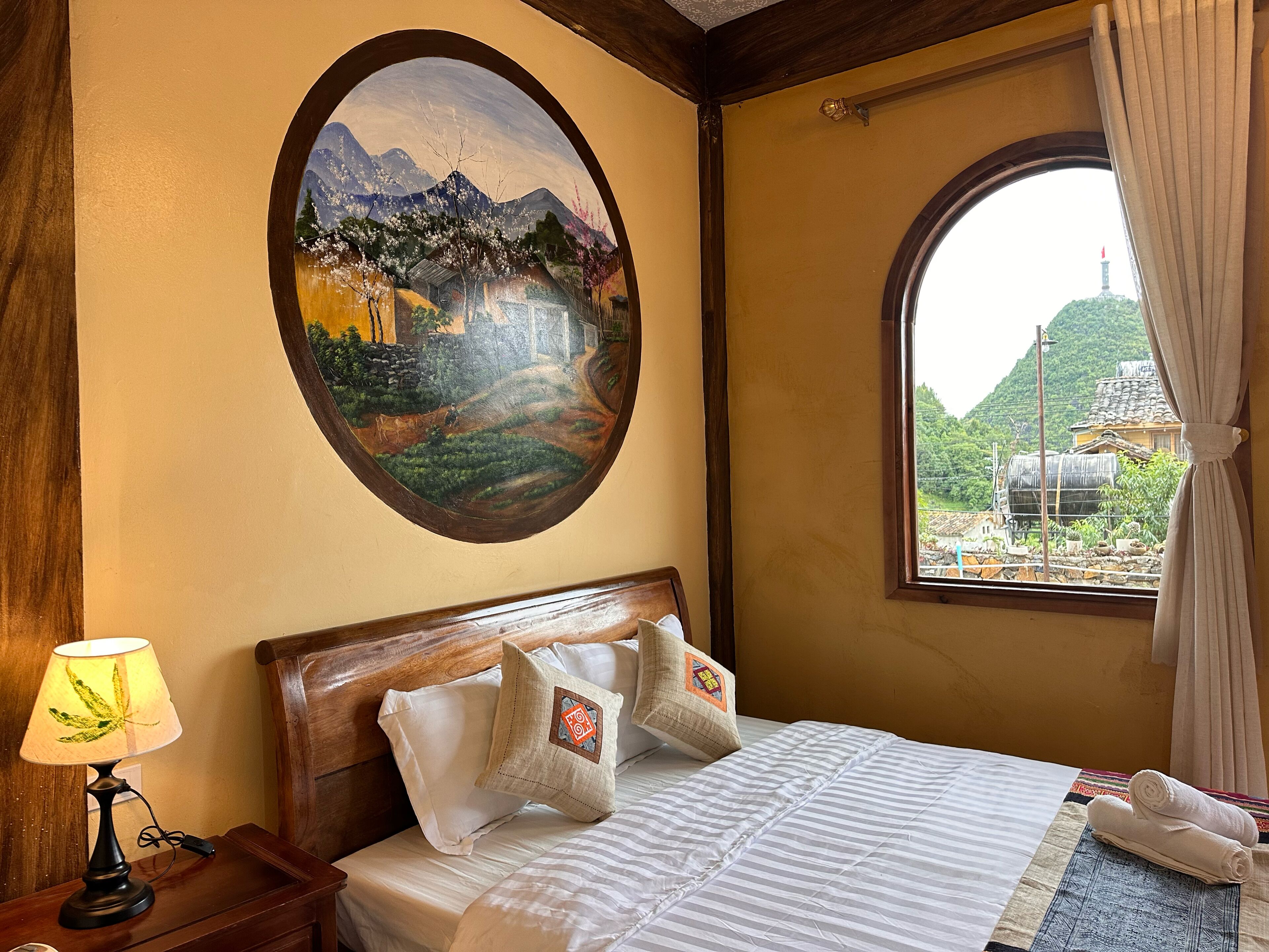 Een hotelkamer met een bed, nachtkastje, lamp, schilderij van een landschap, en een raam met uitzicht op een berg en een gebouw.