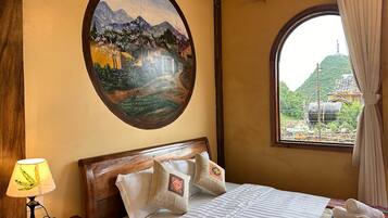 Een hotelkamer met een bed, nachtkastje, lamp, schilderij van een landschap, en een raam met uitzicht op een berg en een gebouw.