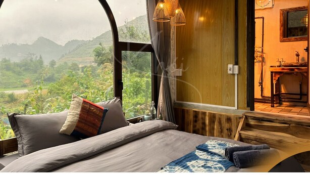 A bedroom with a large window, a bed with pillows, a wooden wall, and a small bathroom visible through the window.