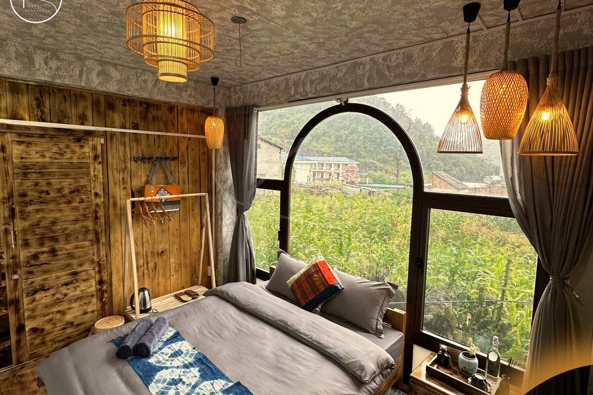 A bedroom with a large window, wooden walls, and a bed with pillows and a blanket.