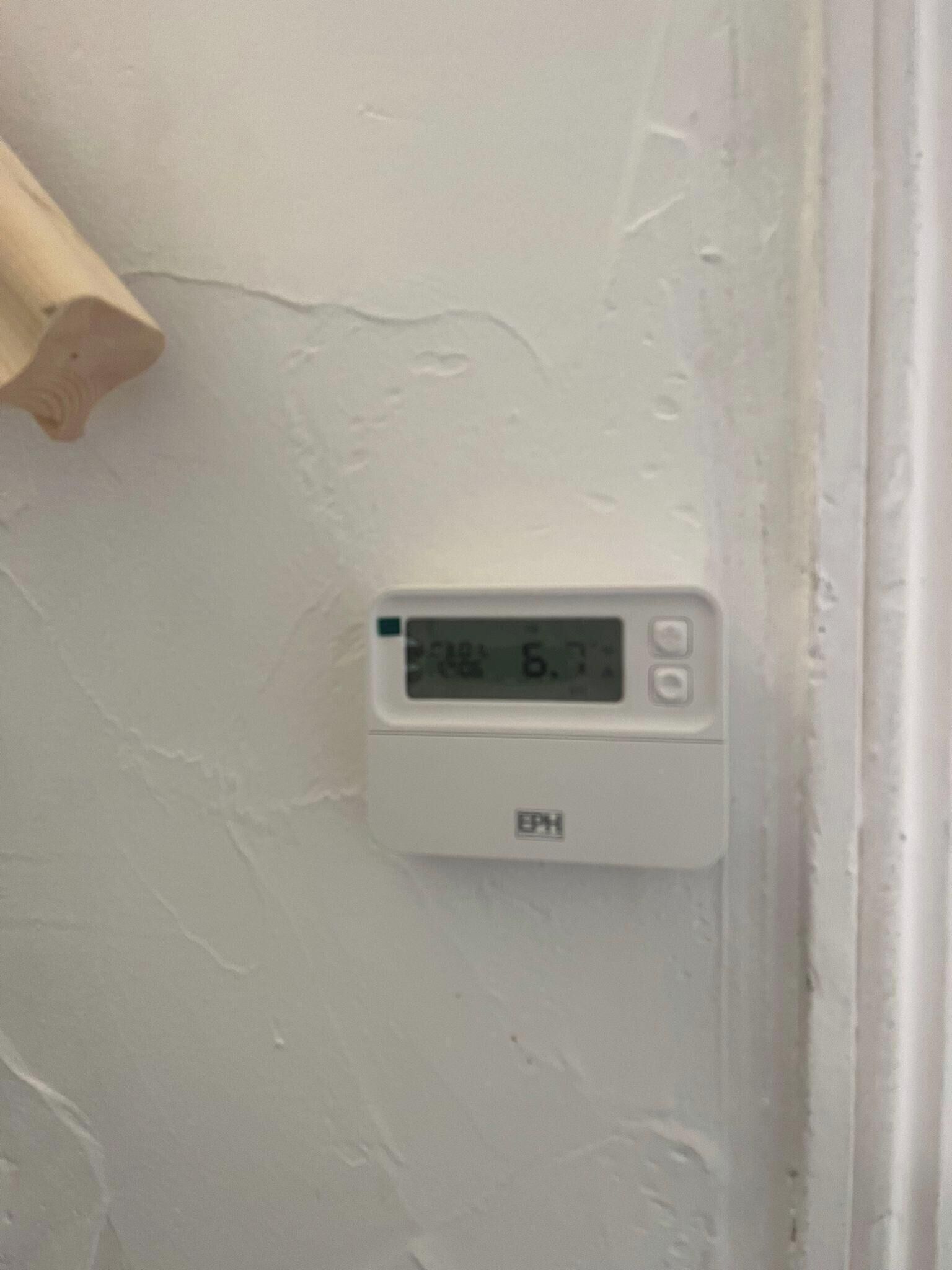 A thermostat mounted on a white wall with a digital display showing the temperature.
