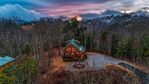 A cabin with a green roof surrounded by trees, a gravel driveway, and a fire pit. - Firefly Ridge! Cozy, Fishing Pond, Hot Tub, Fire Pit! (Sevierville)