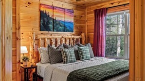 A wooden cabin bedroom with a bed, pillows, a painting, a window, and a nightstand. - Firefly Ridge! Cozy, Fishing Pond, Hot Tub, Fire Pit! (Sevierville)