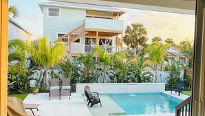 Un espace piscine offrant une vue sur un bâtiment à deux étages, agrémenté de mobilier d’extérieur et d’une végétation luxuriante.