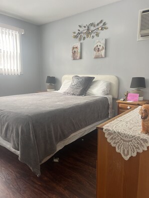 Un dormitorio con cama, dos mesitas de noche, una mesita auxiliar, un adorno en la pared y una ventana con persianas.
