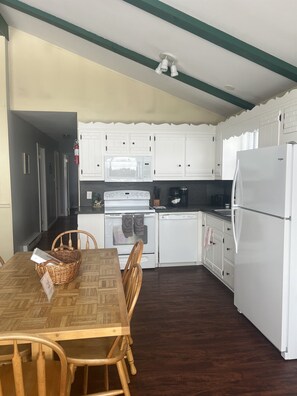A compact kitchen with white cabinets, a microwave, a stove, and a refrigerator. There is a dining table with chairs and a basket.