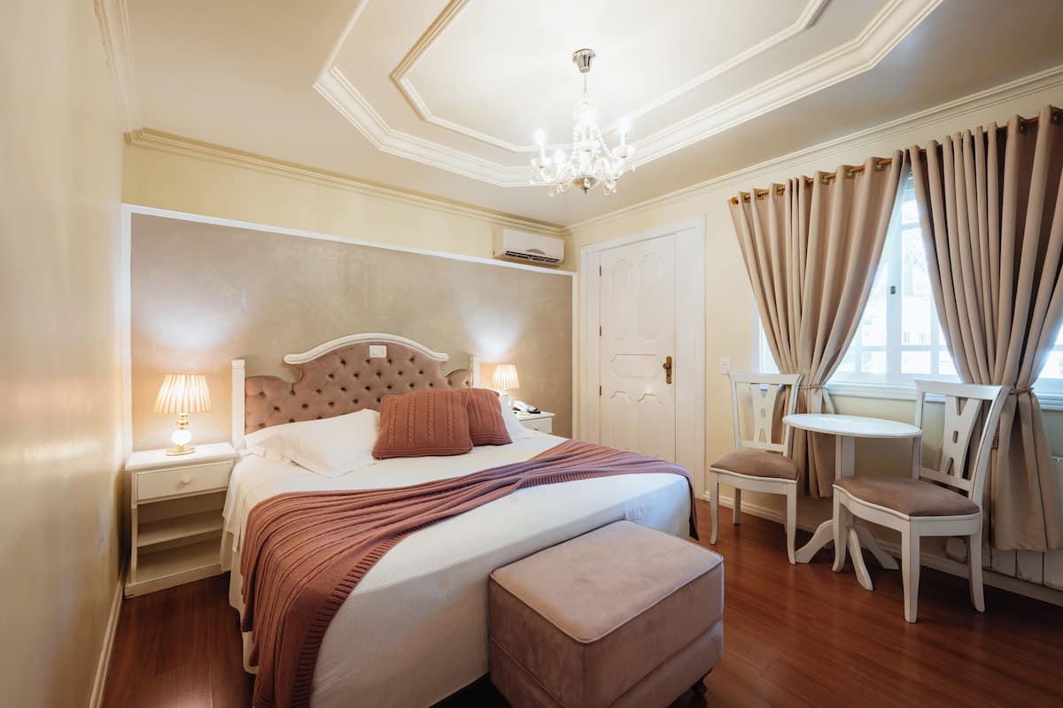 A bedroom with a large bed, bedside tables, a nightstand, a chair, a small table, and a window with curtains.