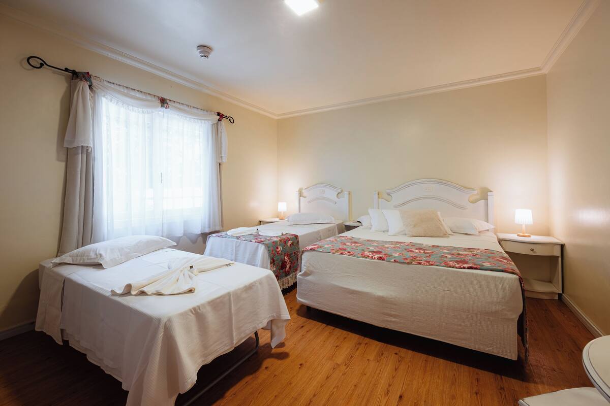 A hotel room with two single beds, a small bedside table with a lamp, a window with curtains, and wooden flooring.