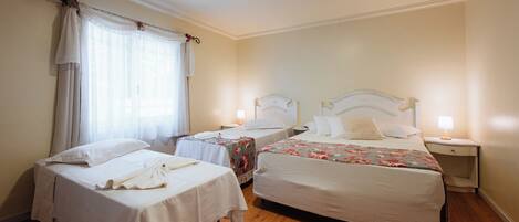 Una habitación de hotel con dos camas individuales, una mesita de noche pequeña con lámpara, una ventana con cortinas y suelo de madera.