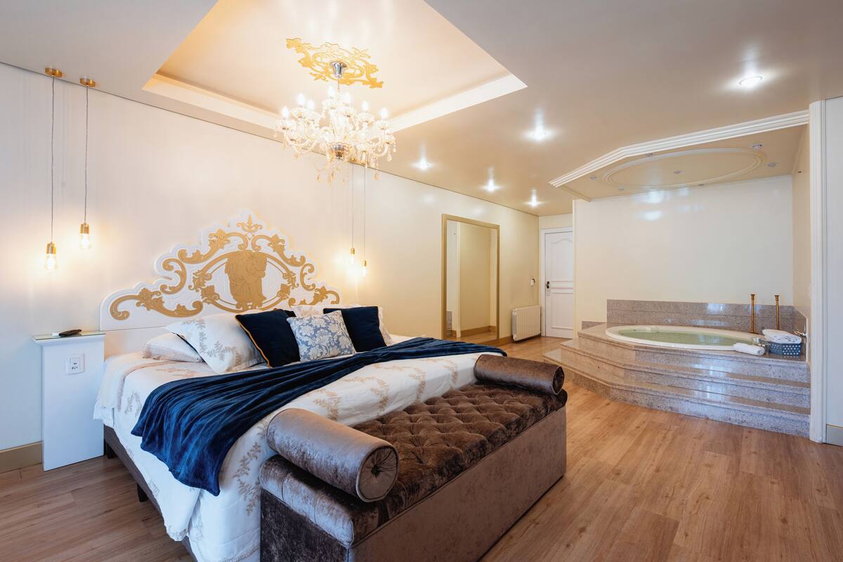 A modern bedroom with a large bed, a chandelier, a built-in bathtub, and a small bedside table.