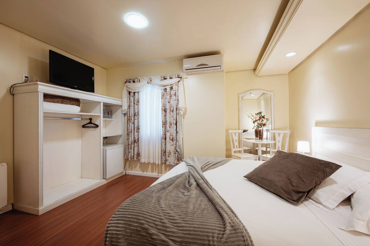 A hotel room with a bed, a TV on a cabinet, a desk with a chair, and an air conditioning unit.