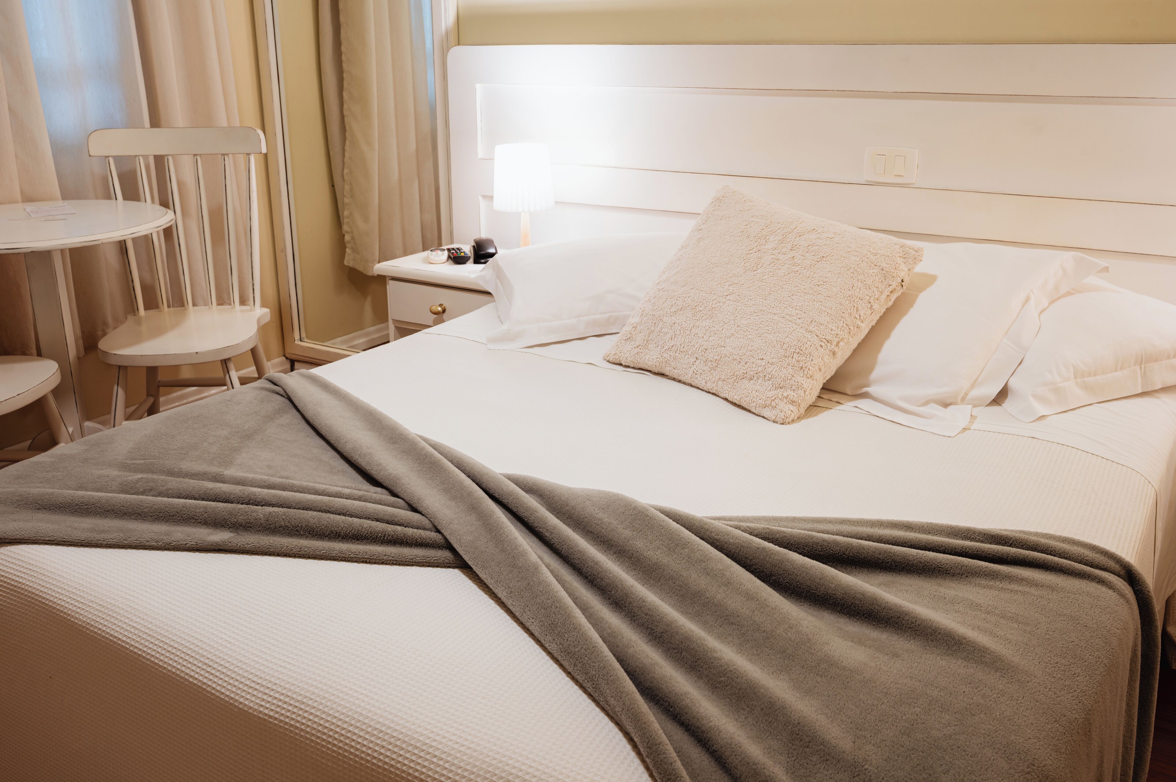 A neatly made bed with a white headboard, a brown throw blanket, and a bedside table with a lamp and a phone.