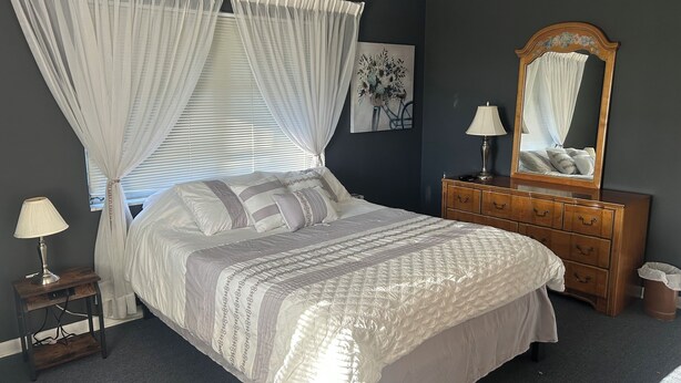 A bedroom with a bed, bedside lamps, a dresser, a mirror, and a painting on the wall. - Charming 3-bedroom house with lake views in serene Spring Hill (Spring Hill)