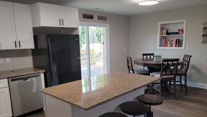 A modern kitchen with white cabinets, a black refrigerator, a dishwasher, and a granite countertop. There is a dining area with a table and chairs, and a bookshelf with books and decorative items. -  Lake Tillery Area, 4-bedroom house in charming Norwood with WiFi, Deck and AC (Norwood)