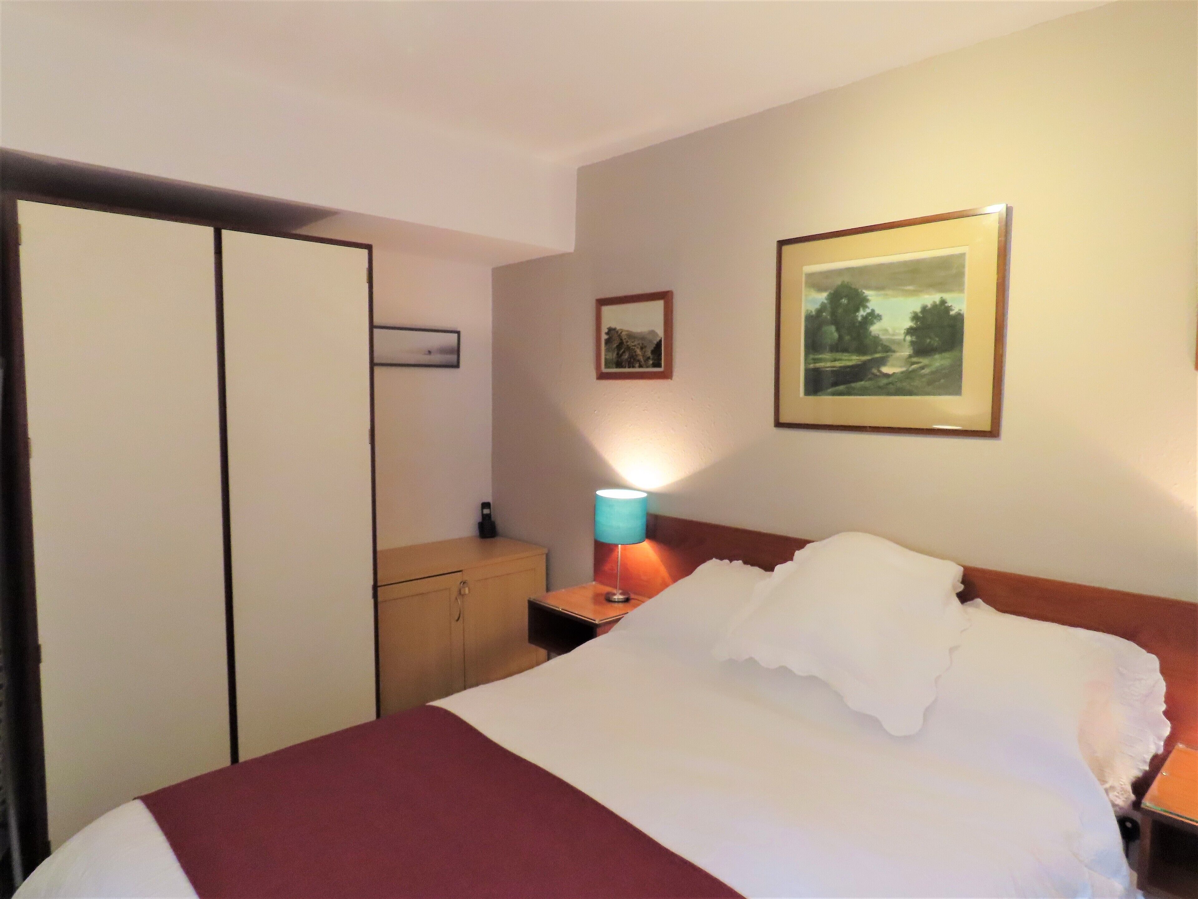 A hotel room with a bed, a nightstand, a lamp, a wardrobe, and two framed pictures on the wall.