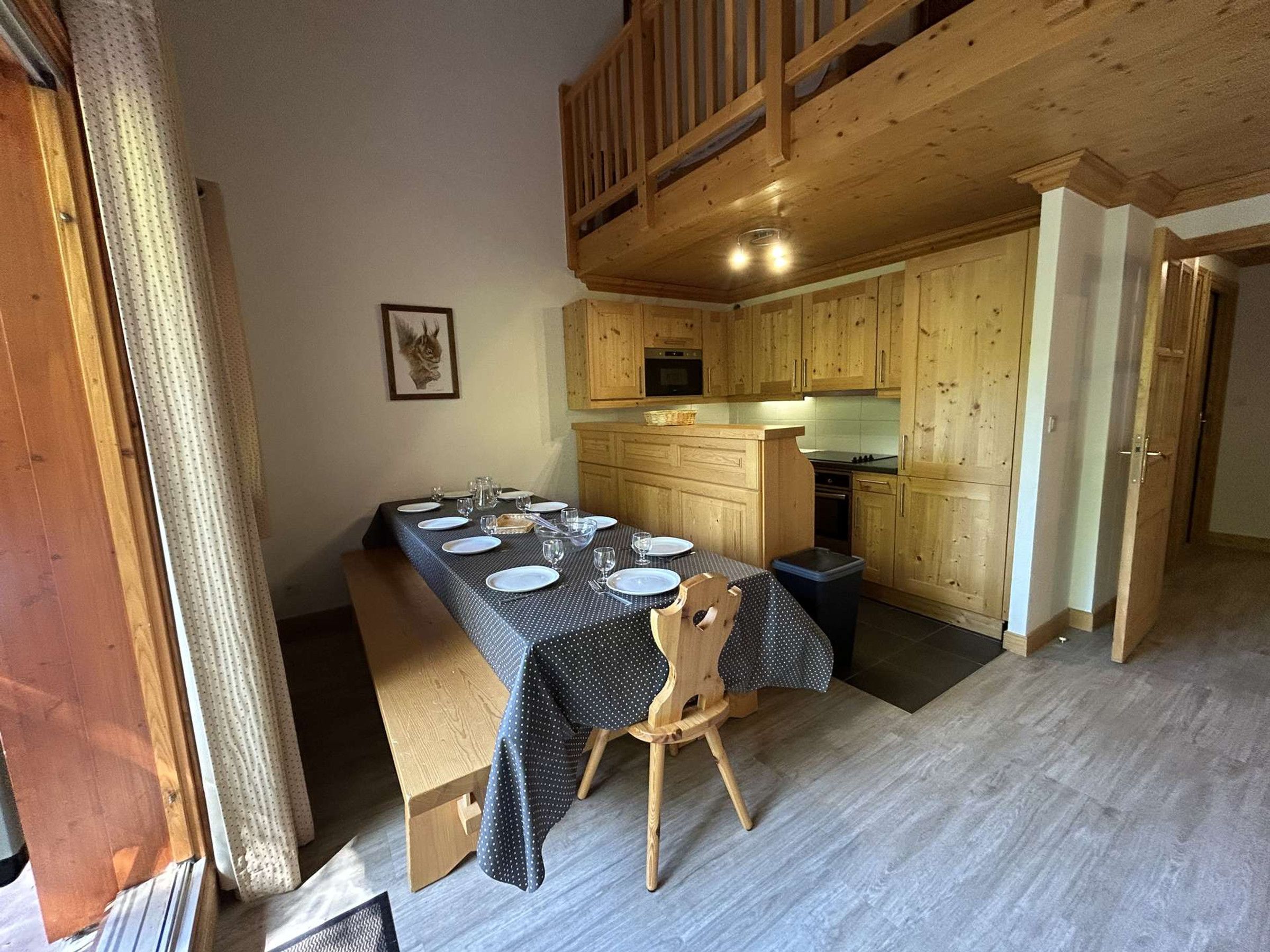 A dining area with a long table set for four, wooden chairs, and a kitchen in the background.