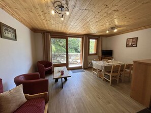 Quarto aconchegante com teto de madeira, mesa de jantar posta para quatro pessoas, televisão e uma grande janela com vista para o verde.