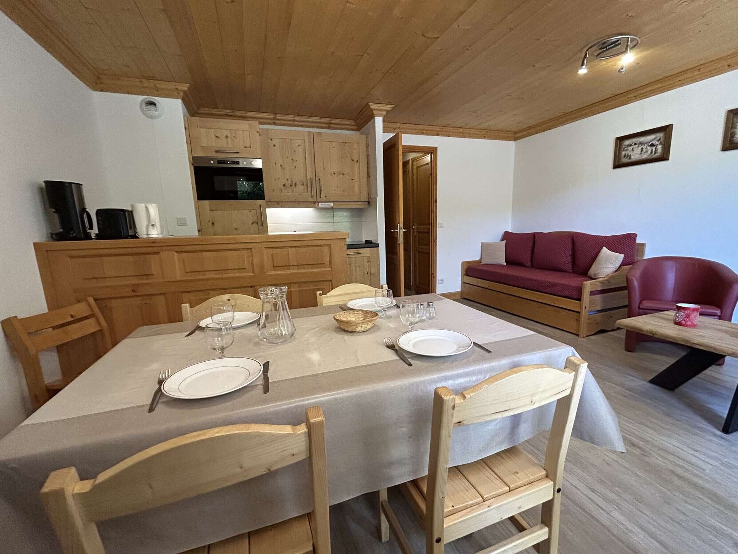 Uma sala de jantar com uma mesa posta para quatro pessoas, cadeiras de madeira, uma cozinha com armários de madeira, um sofá vermelho e uma poltrona vermelha.