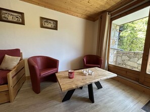 Quarto com teto de madeira, mesa de madeira com pés pretos, duas cadeiras vermelhas, sofá de madeira com almofada vermelha e parede de pedra visível pela janela.