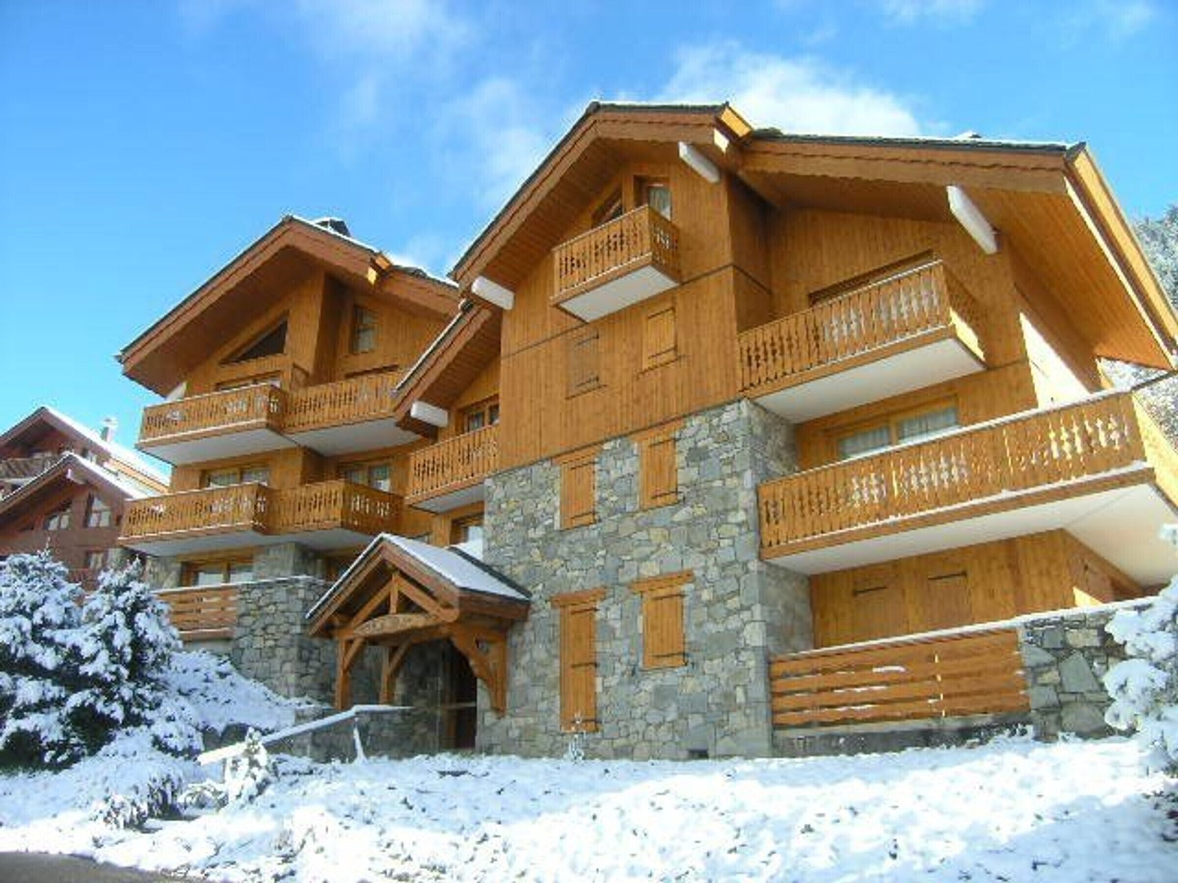 A multi-story wooden chalet with balconies, stone walls, and a snow-covered landscape.
