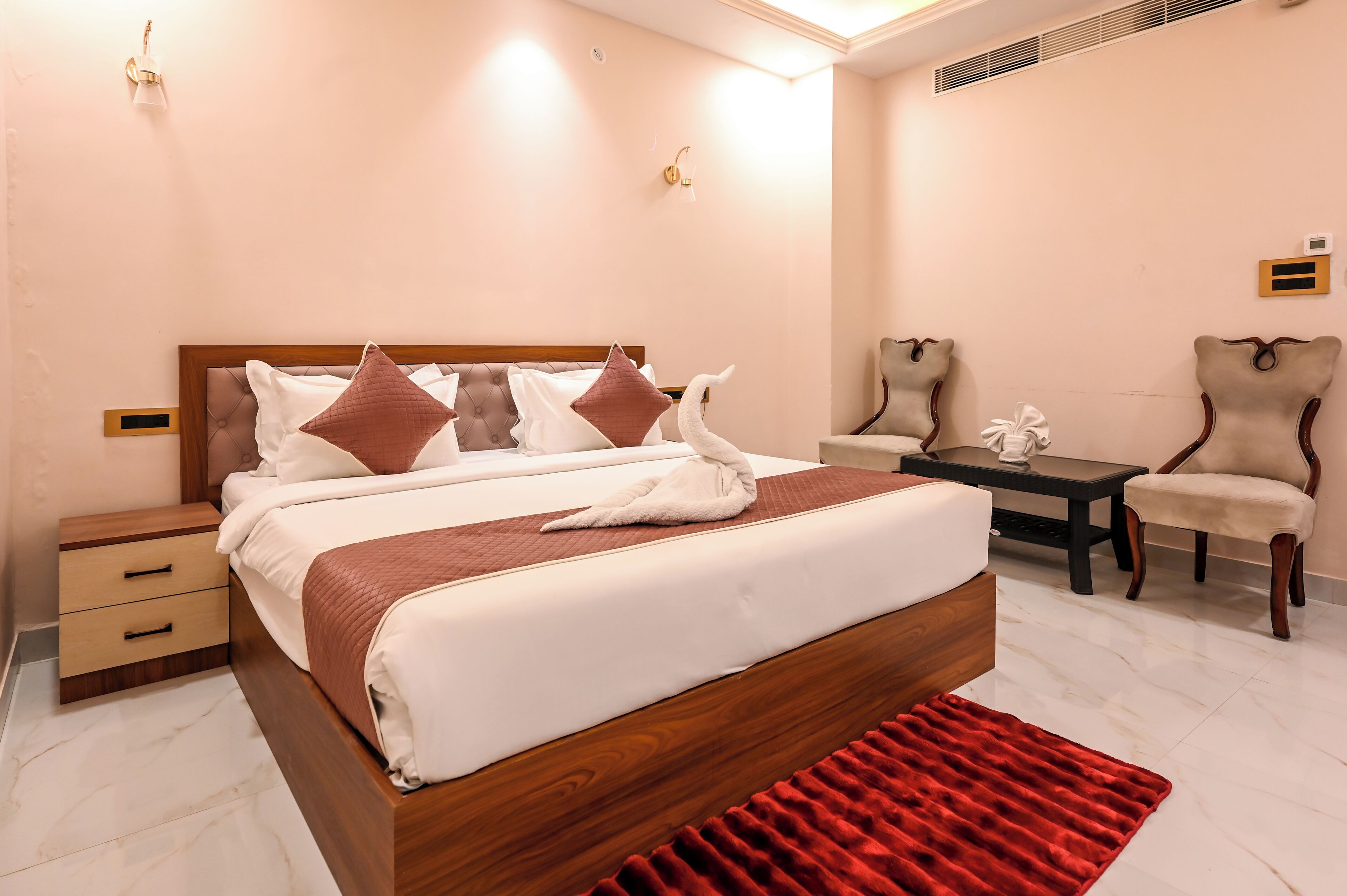 Una cama bien hecha con un edredón rojo, una mesita de noche de madera con cajones, una alfombra roja, una pequeña mesa con una estatua blanca y dos sillones.
