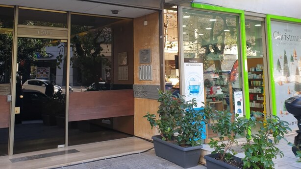 Fachada con un letrero verde, amplias ventanas y plantas en macetas al frente.