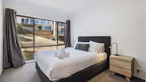 A modern bedroom with a large bed, a nightstand, and a view of a building through floor-to-ceiling windows. - Kirwan 8, 2 bedroom apartment (Jindabyne)