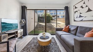 A modern living room with a grey sofa, a coffee table, a TV, and a large window offering a view of a brick building and greenery. - Kirwan 8, 2 bedroom apartment (Jindabyne)