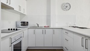A modern kitchen with white cabinetry, a microwave, a sink, and a clock on the wall. - Kirwan 8, 2 bedroom apartment (Jindabyne)