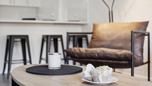 A modern living room with a brown leather sofa, a round wooden coffee table, and a white plate with pastries. - Kirwan 8, 2 bedroom apartment (Jindabyne)