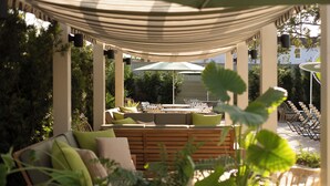 A covered outdoor seating area with a wooden bench, cushioned chairs, and a large umbrella. - The Maidstone New Orleans (New Orleans)