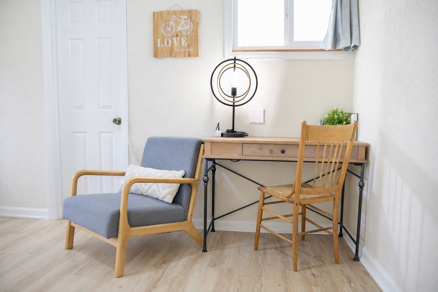 Een kamer met een bureau, twee stoelen, een lamp, een raam met gordijn en een deur.