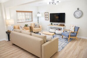 A living room with a sofa, two armchairs, a coffee table, a TV, a clock, and a window with blinds.