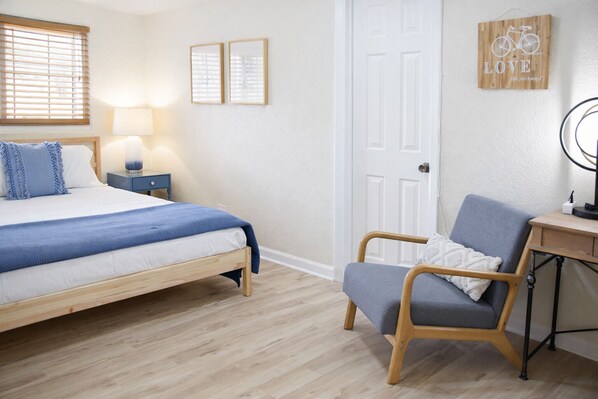 A bedroom with a bed, bedside table, lamp, chair, desk, and door.