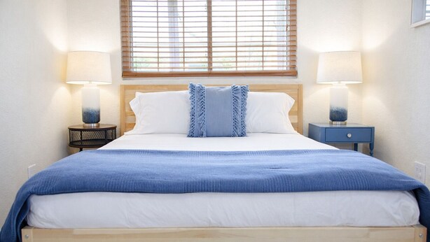 Un lit avec un cadre en bois, une literie blanche et une couverture bleue. Il y a deux tables de chevet avec des lampes, une fenêtre avec des stores et une étagère fixée au mur.