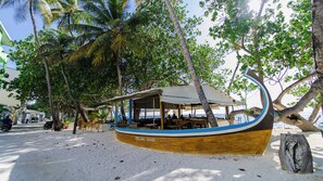 Ein Boot mit überdachter Sitzgruppe, benannt 'VELAA VANI', liegt an einem sandigen Strand mit Palmen und blauem Himmel.