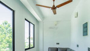 A modern living room with a ceiling fan, large windows, a grey sofa, and a small wooden table. - VelaaVani - Turtle Hive Dhiffushi (Dhiffushi)