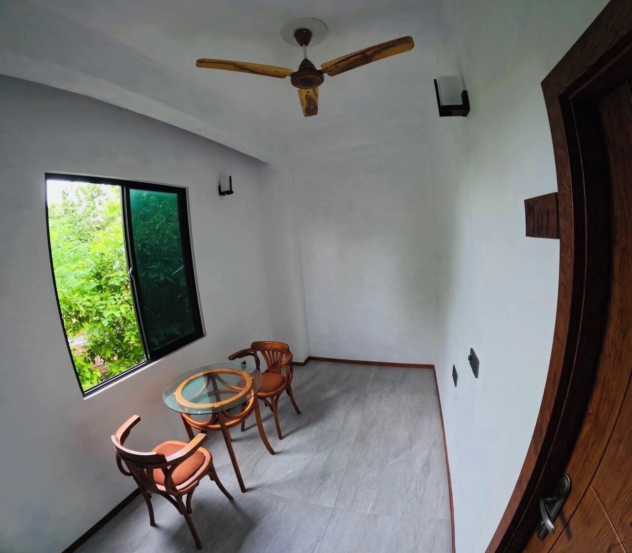 A room with a ceiling fan, a window with a view of greenery, and a dining area with a table and chairs.
