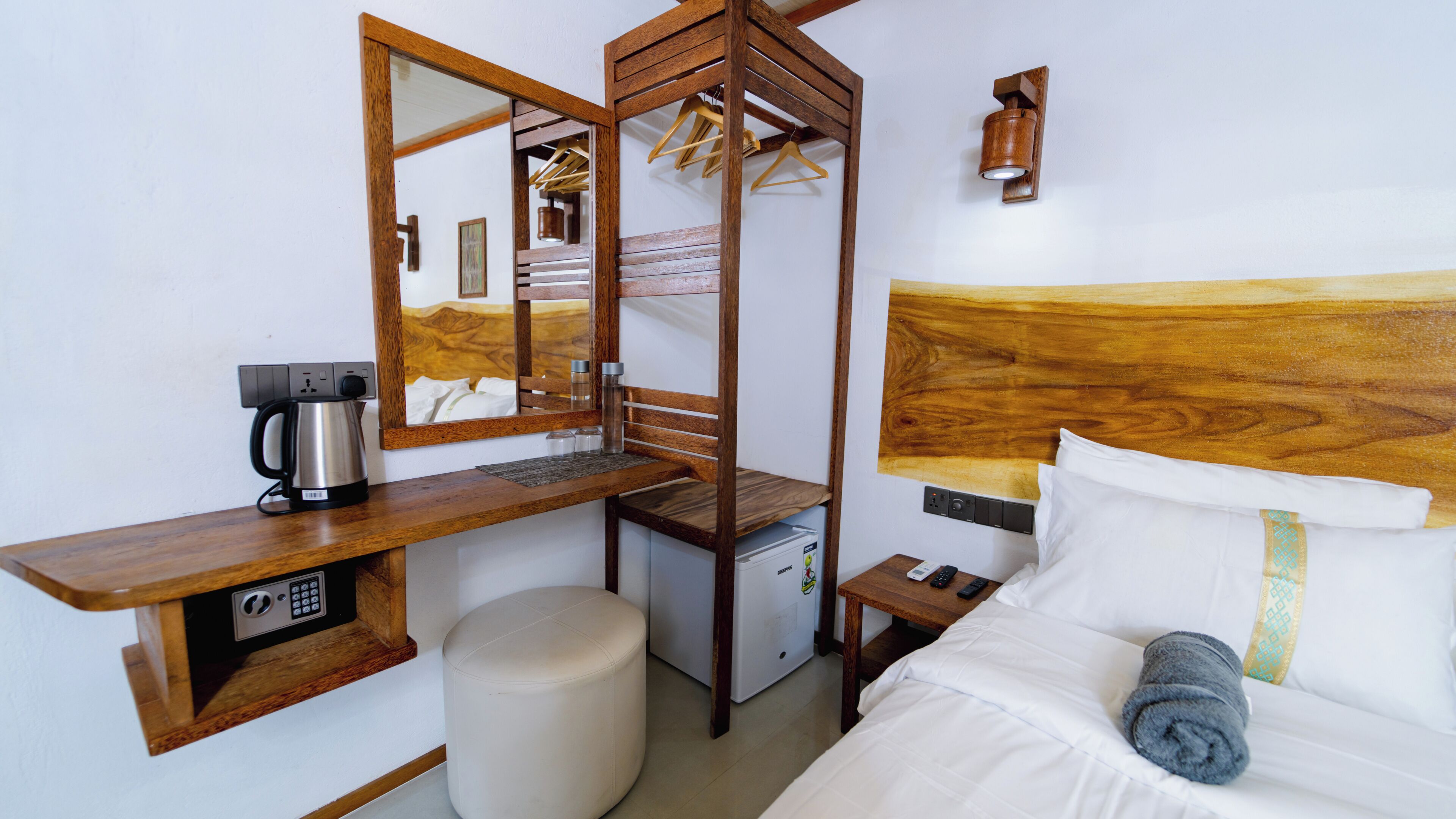A hotel room with a wooden desk, a bed with white linens, a bedside table with a remote control, a coffee maker, and a mirror.