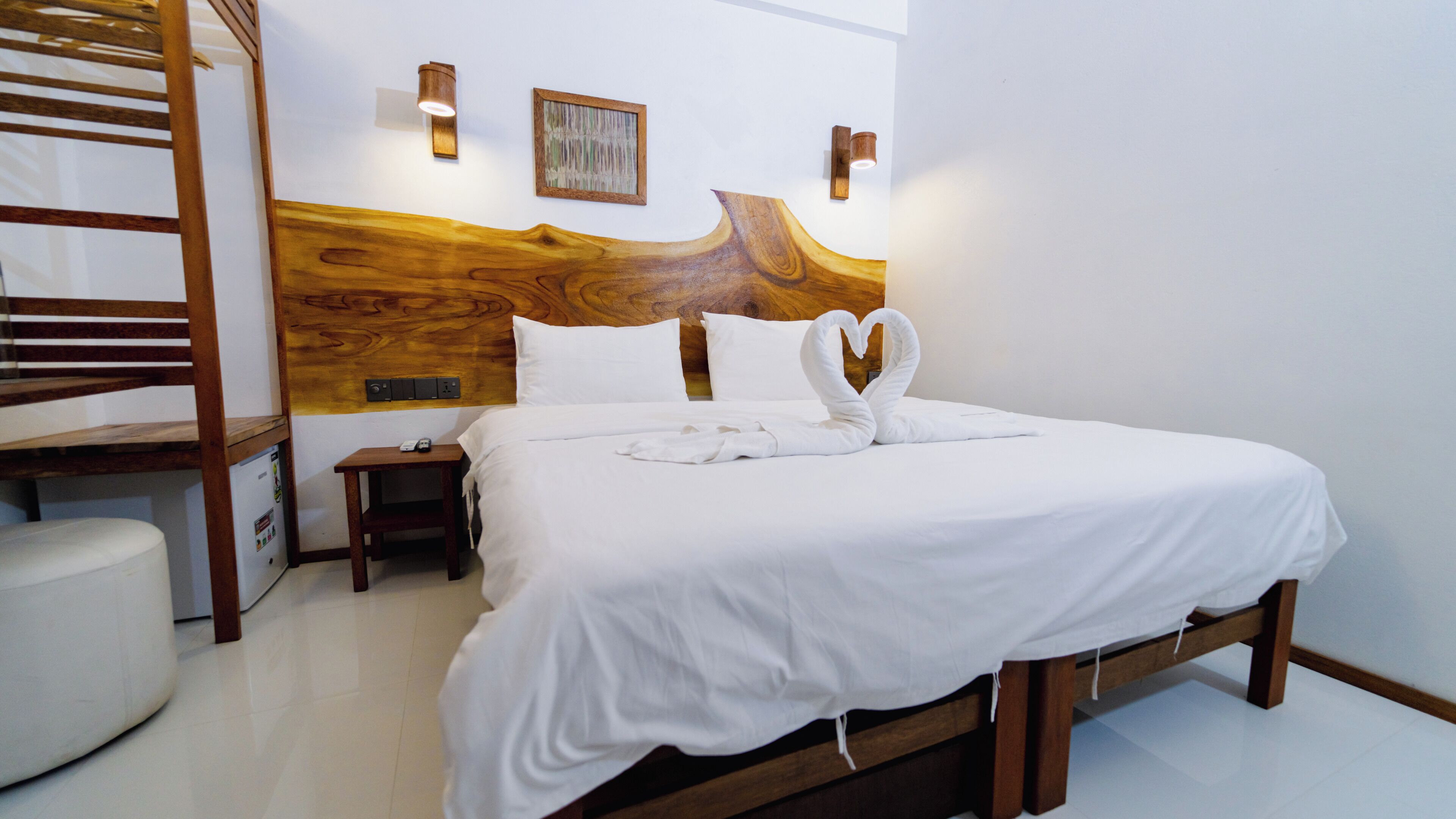 Habitación de hotel con cabecera de madera, cama con ropa blanca, mesita de noche con control remoto y lámpara de pared.