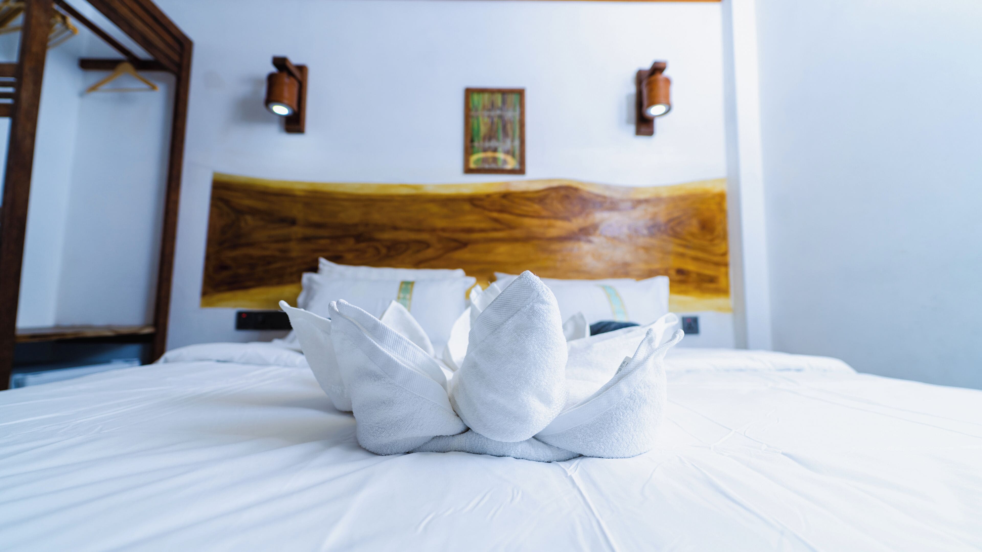 A neatly made bed with a towel arrangement in the center, a wooden headboard, and two wall-mounted lights.