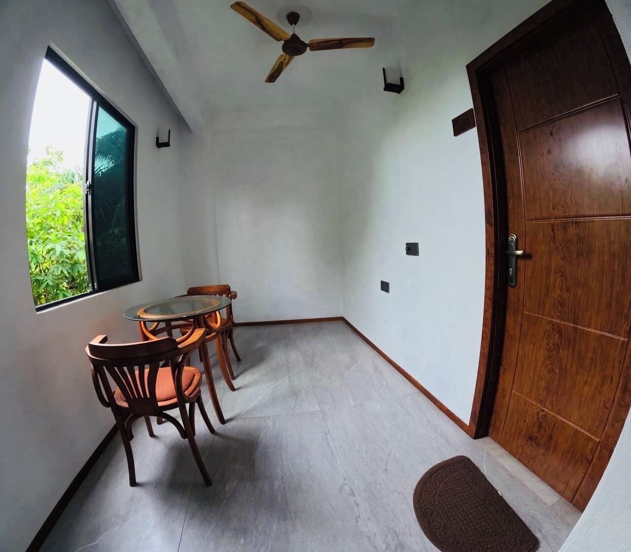 Una habitación con puerta de madera, ventilador de techo, mesa de comedor con sillas y ventana con vistas a follaje.