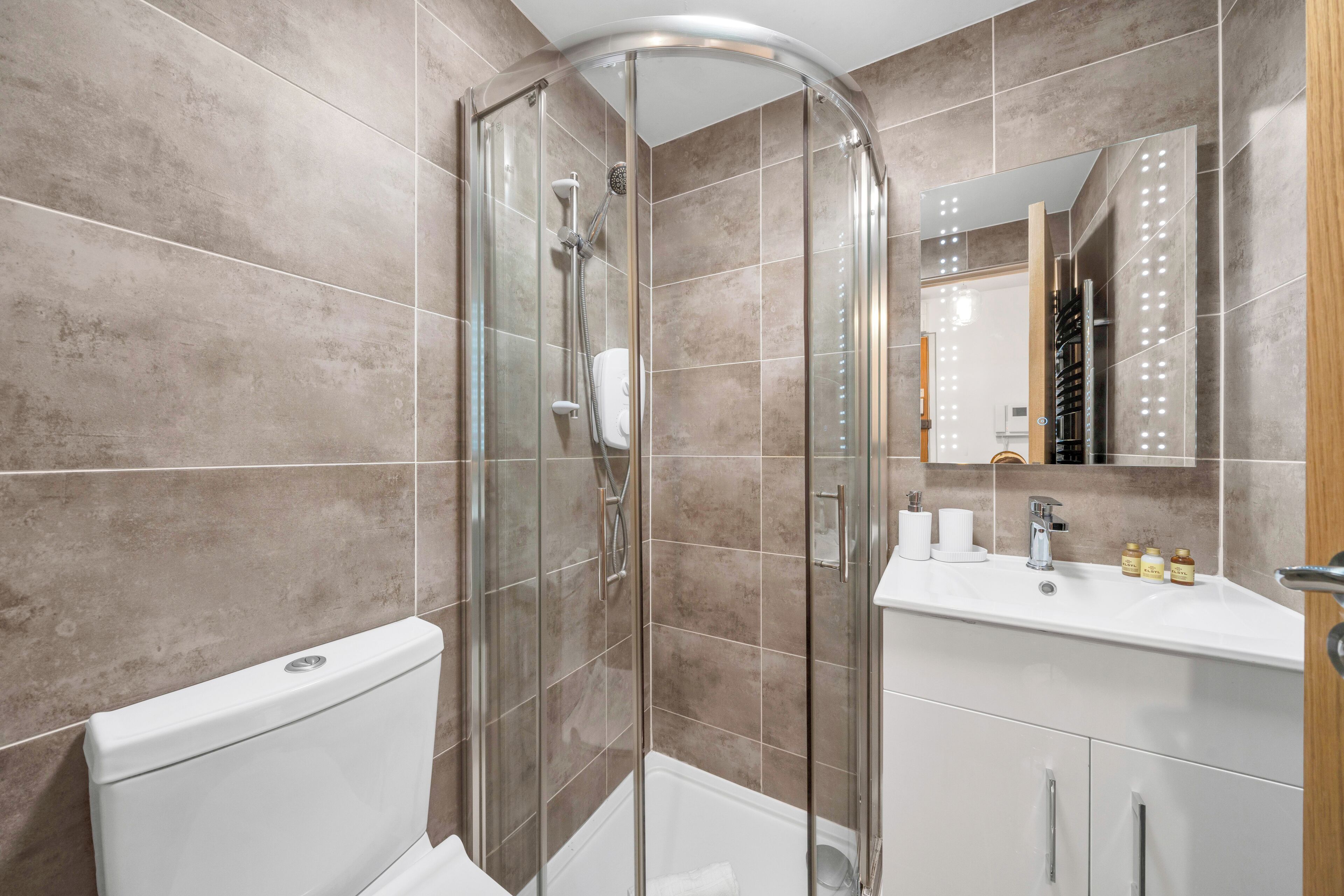 A modern bathroom with a glass shower enclosure, a white toilet, a sink with toiletries, and a mirror with lights.