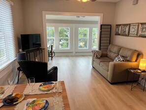 A living room with a sofa, a television, a dining table with plates and a glass, and a view through a glass door to the outside. - Casa Carrera 1 BR w/ Sunroom-Easy Walk to Town (St. Augustine)