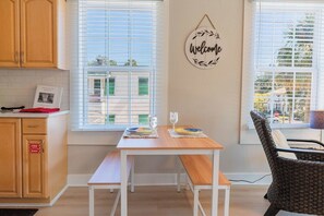 A dining area with a wooden table and benches, a wicker chair, and a wall-mounted welcome sign. - Casa Carrera 1 BR w/ Sunroom-Easy Walk to Town (St. Augustine)