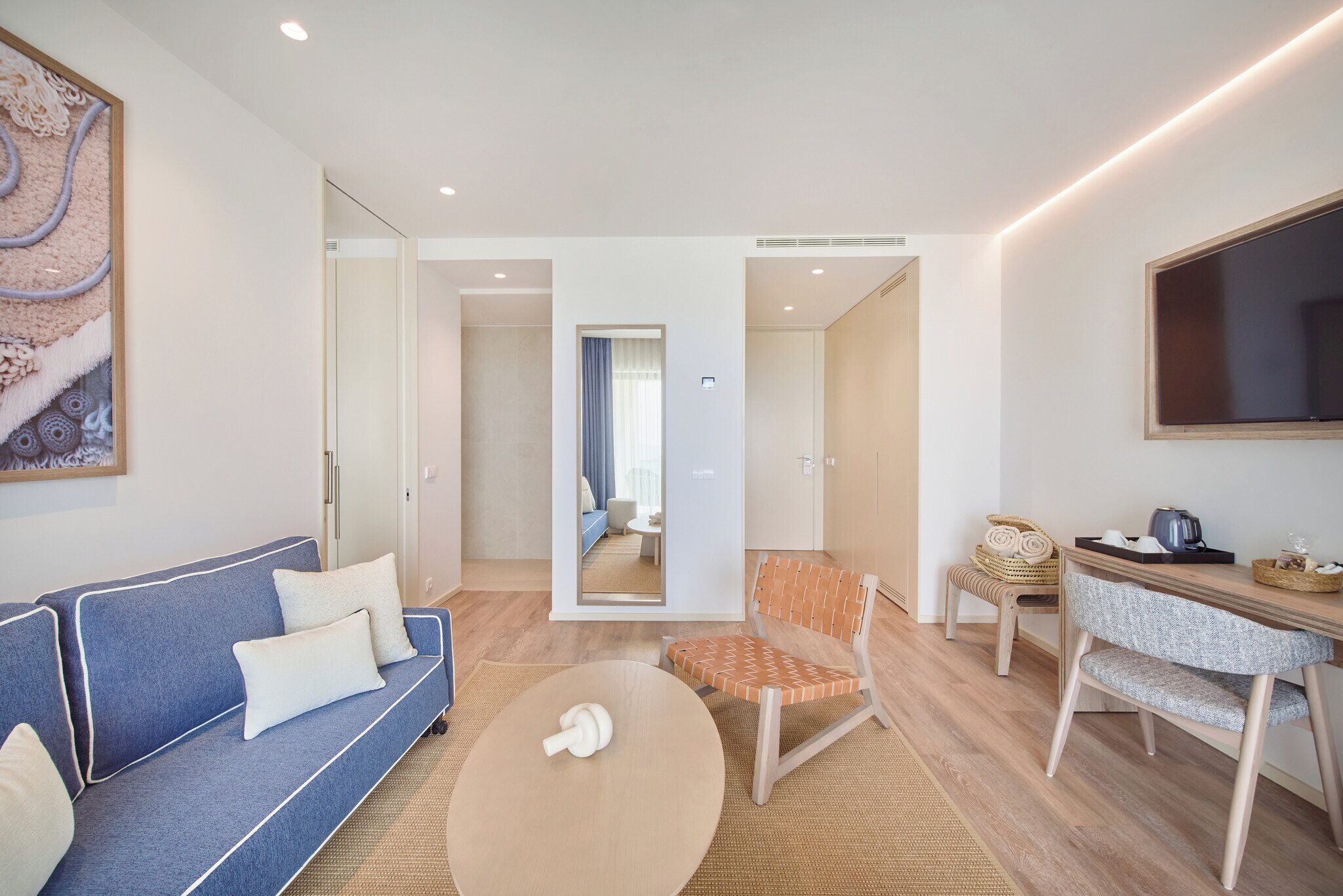 A modern hotel room with a blue sofa, a wooden chair, a small table, and a large mirror.