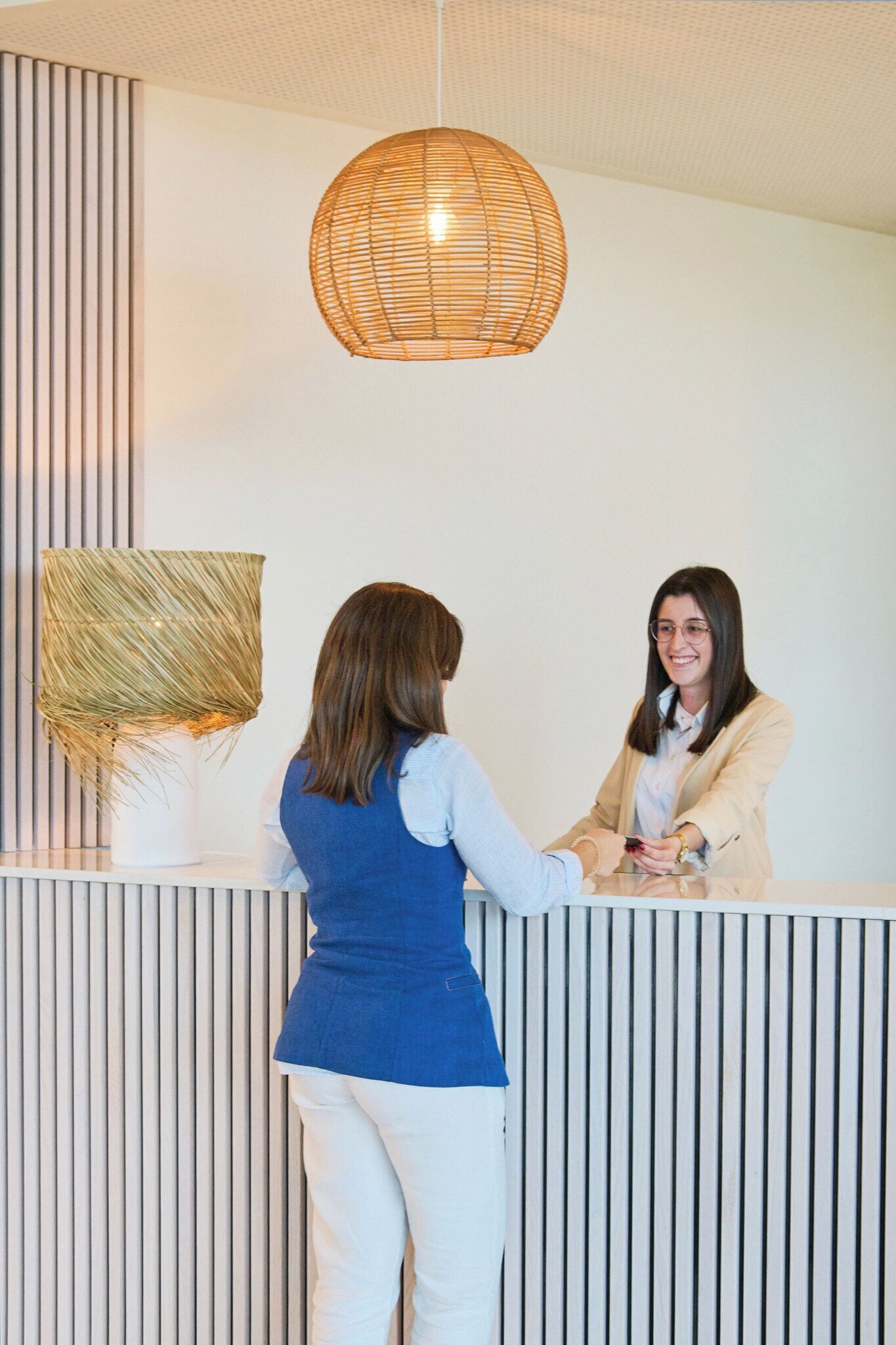 A reception area with a modern design, featuring a large woven pendant light, a white reception desk, and vertical paneling on the wall.