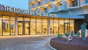 Ett hotell med en modern fasad som kännetecknas av geometriska mönster och balkonger. Namnet ”THE EDITORY BY THE SEA HOTEL” syns ovanför entrén.