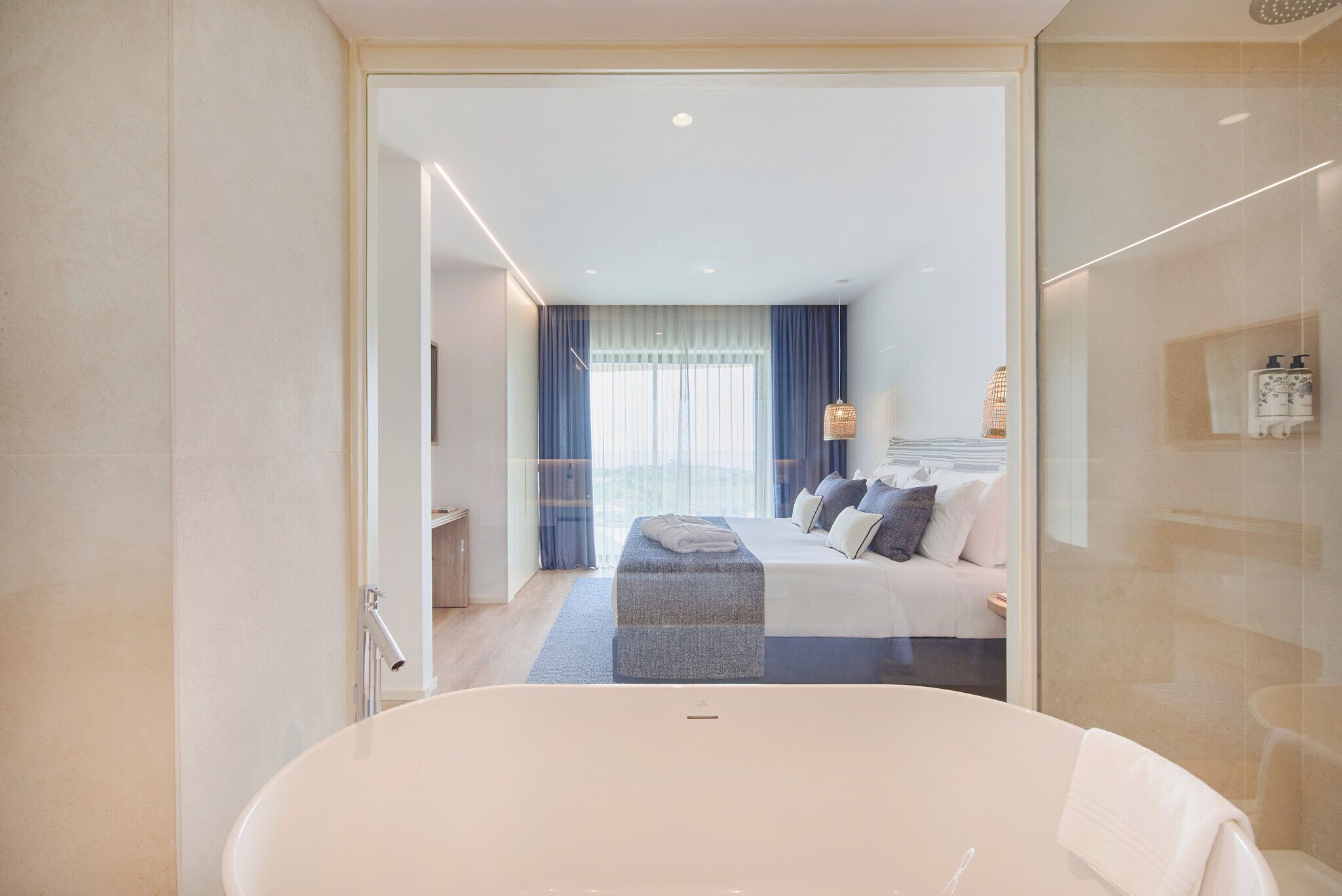 Ein modernes Hotelzimmer mit einer großen Badewanne, einem ordentlich bezogenen Bett und Blick auf die Stadt durch große Fenster.