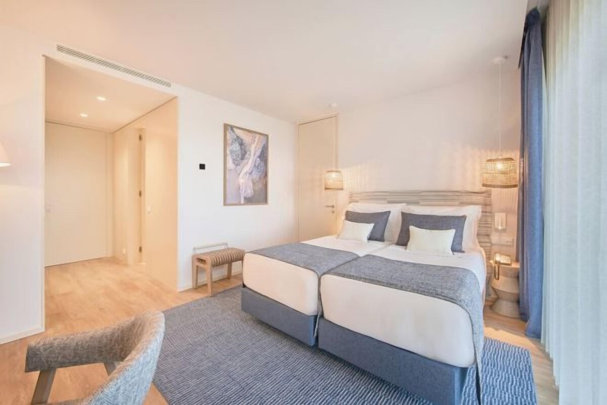 A modern hotel room with a double bed, a small bedside table, a chair, and a painting on the wall.